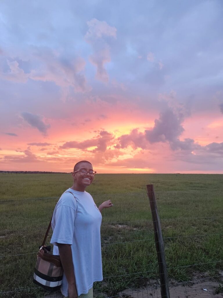 Person in a light blue shirt stands near a grassy field at sunset, smiling and pointing toward the colorful sky with pink and orange clouds.