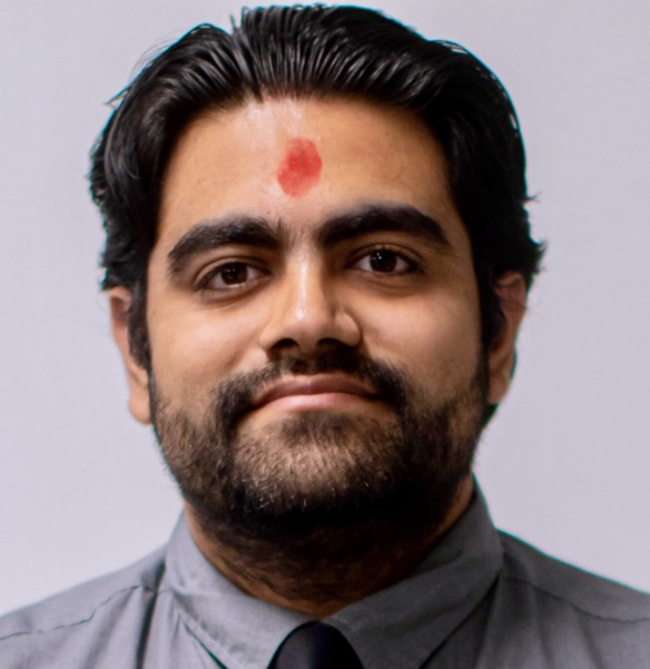A man with a beard and a red mark on his forehead is wearing a gray collared shirt and black tie, posing in front of a plain background.
