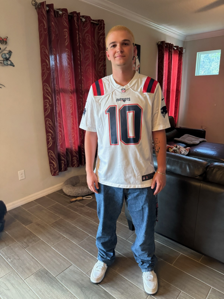 A person with short blond hair stands indoors on tile flooring, wearing a white Patriots football jersey, blue jeans, and white sneakers. Red curtains and a brown sofa are in the background.